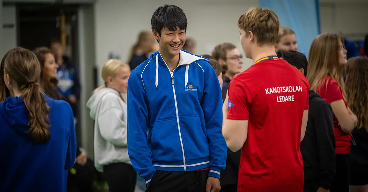 En ung man med en blå tröja leende, omgiven av andra personer i en lokal. En person bär en röd tröja med texten ”KANOTSKOLAN LEDARE”.