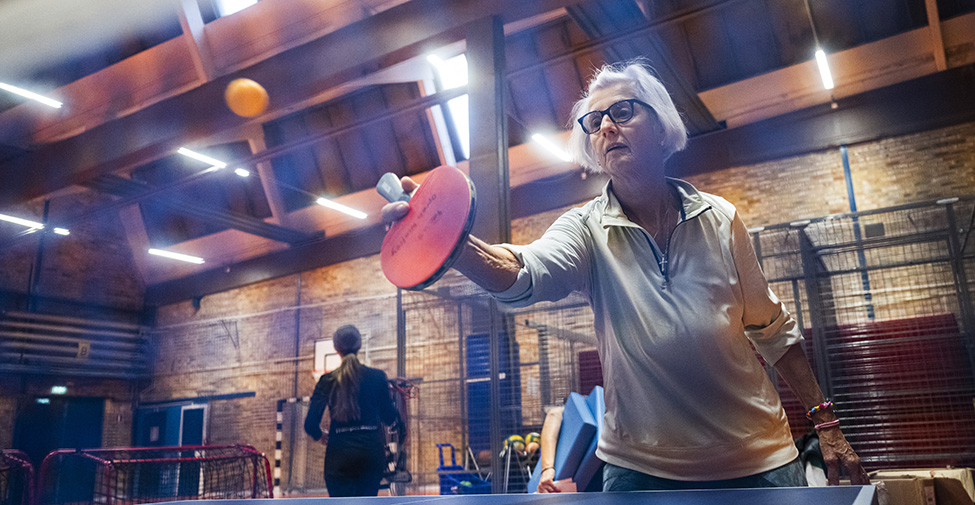 En äldre kvinna spelar bordtennis och slår en boll i luften vid ett pingisbord. I hallen syns också en person i bakgrunden.