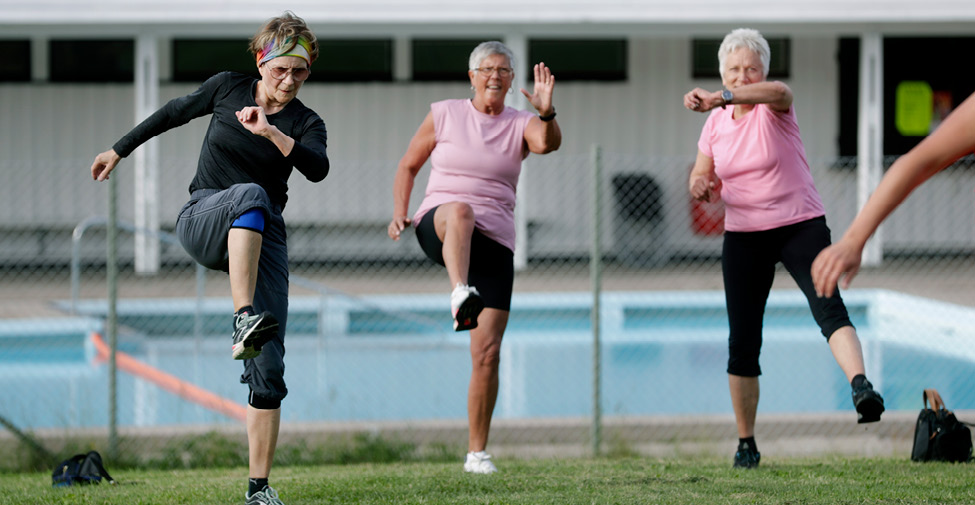 Tre kvinnor i övre medelåldern utför gympa utomhus. 