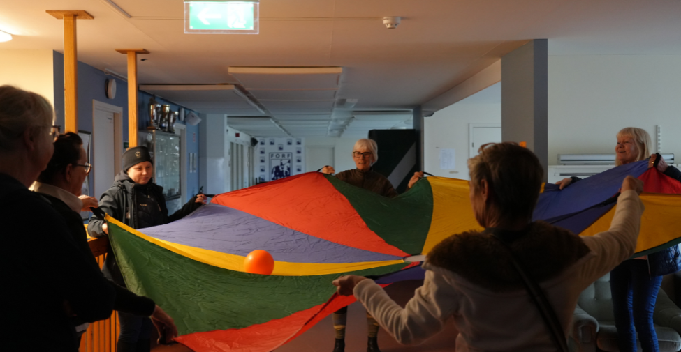 Flera personer håller i en regnbågsfärgat tygstycke, med en orange boll som rullar på tyget.