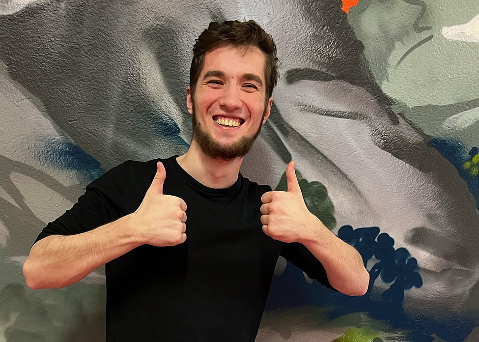 En glad ung man gör två tummar upp framför en färgglad vägg.