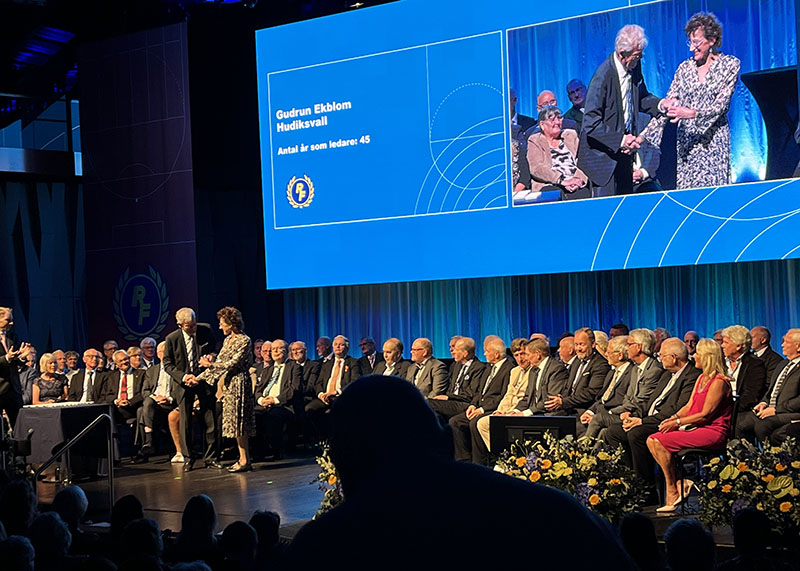 Ett 70-tal personer sitter på en scen, R:s ordförande delar ut guldnålen till Gudrun Ekblom.