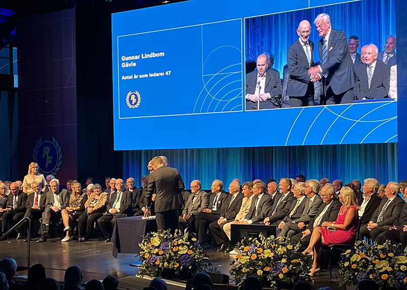 Ett 70-tal personer sitter på en scen, ordförande delar ut guldnålen till Gunnar Lindbom