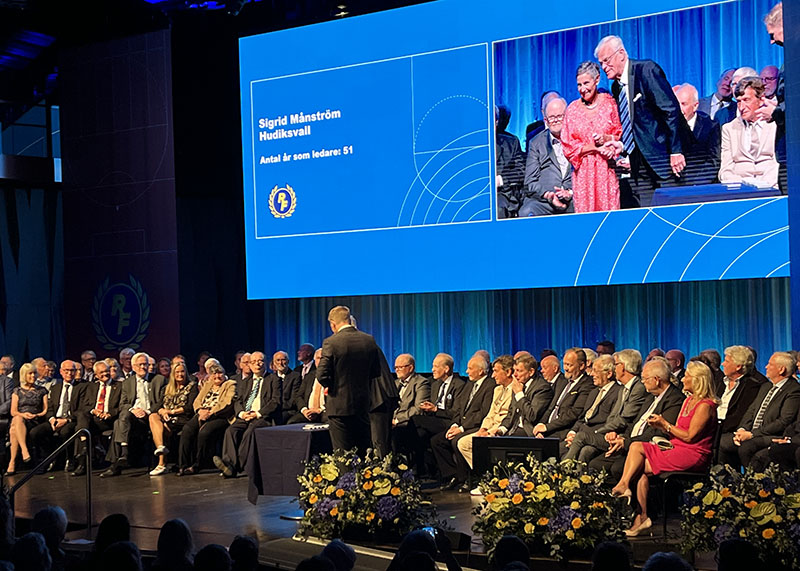 Ett 70-tal personer sitter på en scen, ordförande delar ut guldnålen till Sigrid Månström