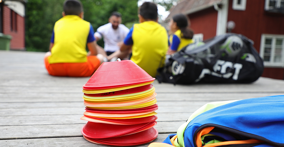 Koner, bollar och annat material för idrottsaktiviteter i fokus, med människor som sitter i bakgrunder i utomhusmiljö